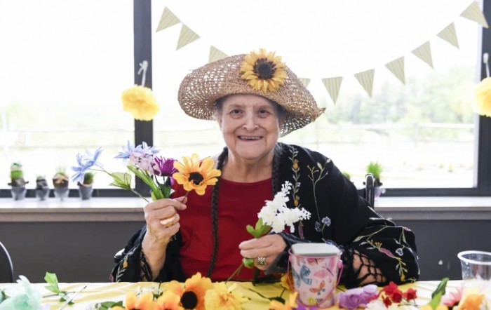 An elderly woman wearing a straw hat adorned with flowers, holding colorful flowers and smiling indoors.