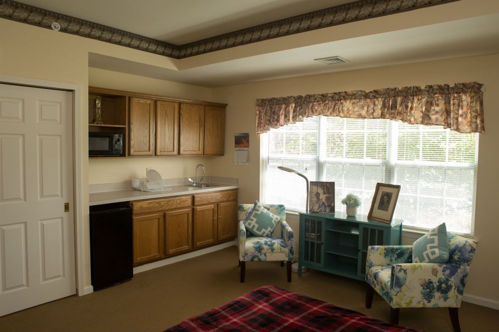 A cozy room with a kitchenette featuring wooden cabinets, two upholstered chairs, and a large window allowing natural light in.