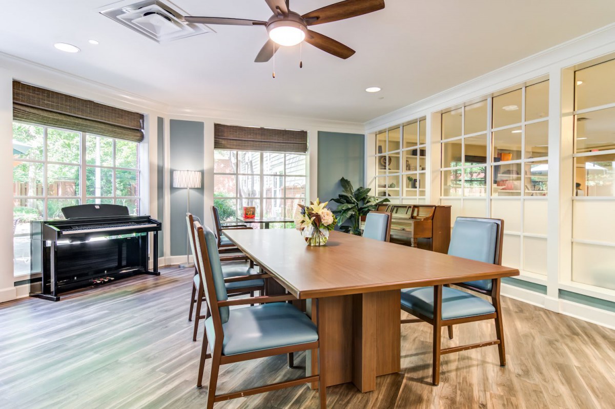 A cozy dining room featuring a large wooden table with chairs, a piano in the corner, and natural light streaming in through the windows.