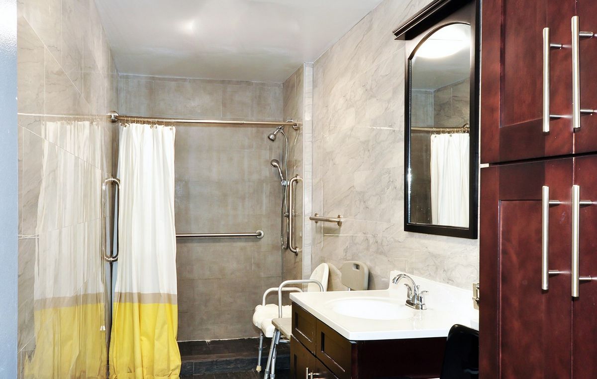 An accessible bathroom featuring a shower with grab bars, a chair, and a sink with a mirror.