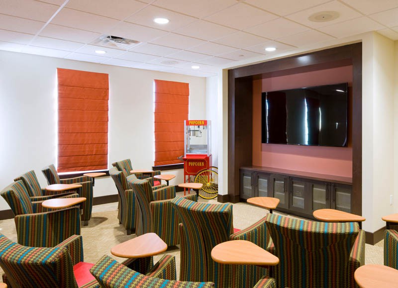 A modern classroom with colorful seating arrangements, a large television, and windows with red blinds.