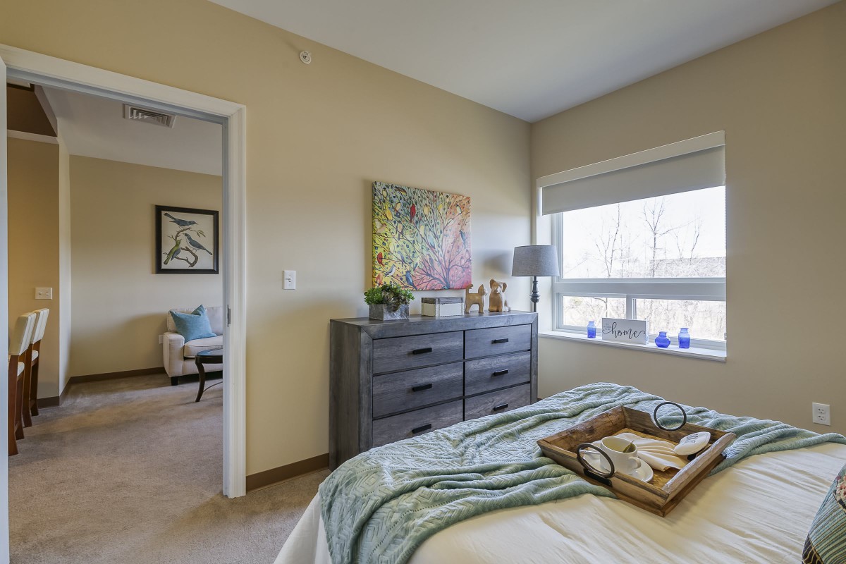 A cozy bedroom with a dresser, artwork, and a large window letting in natural light.