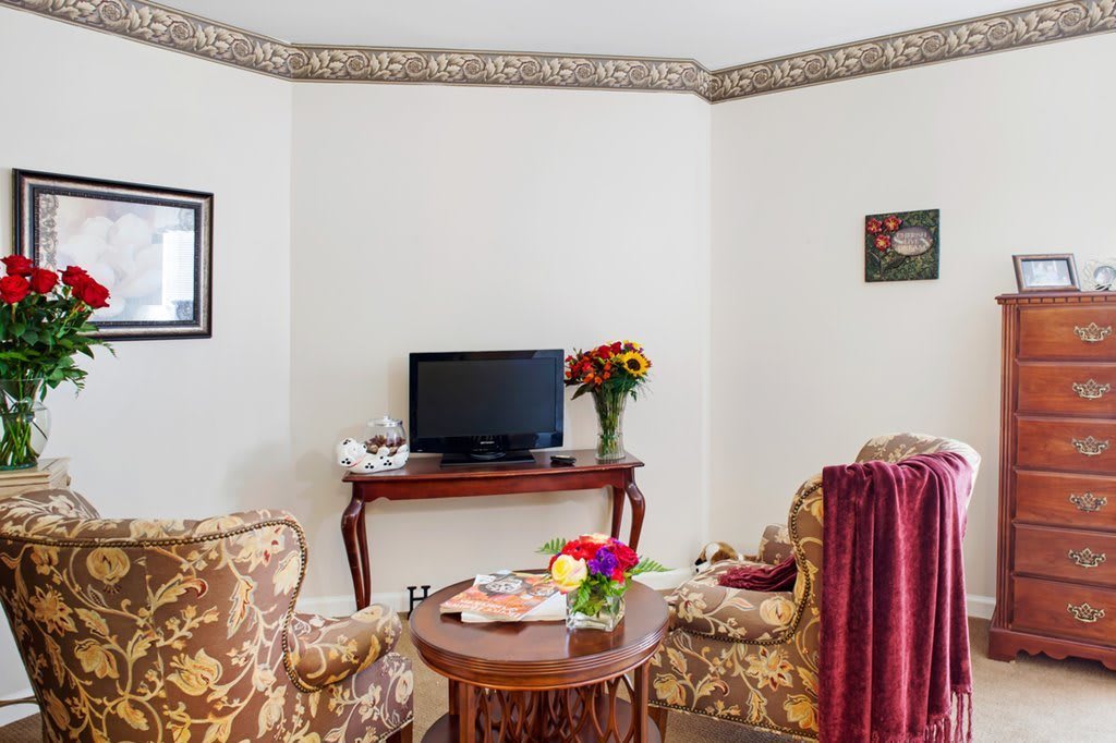 A cozy living room setup featuring elegant chairs, a small table with a bouquet of flowers, and a television on a console.