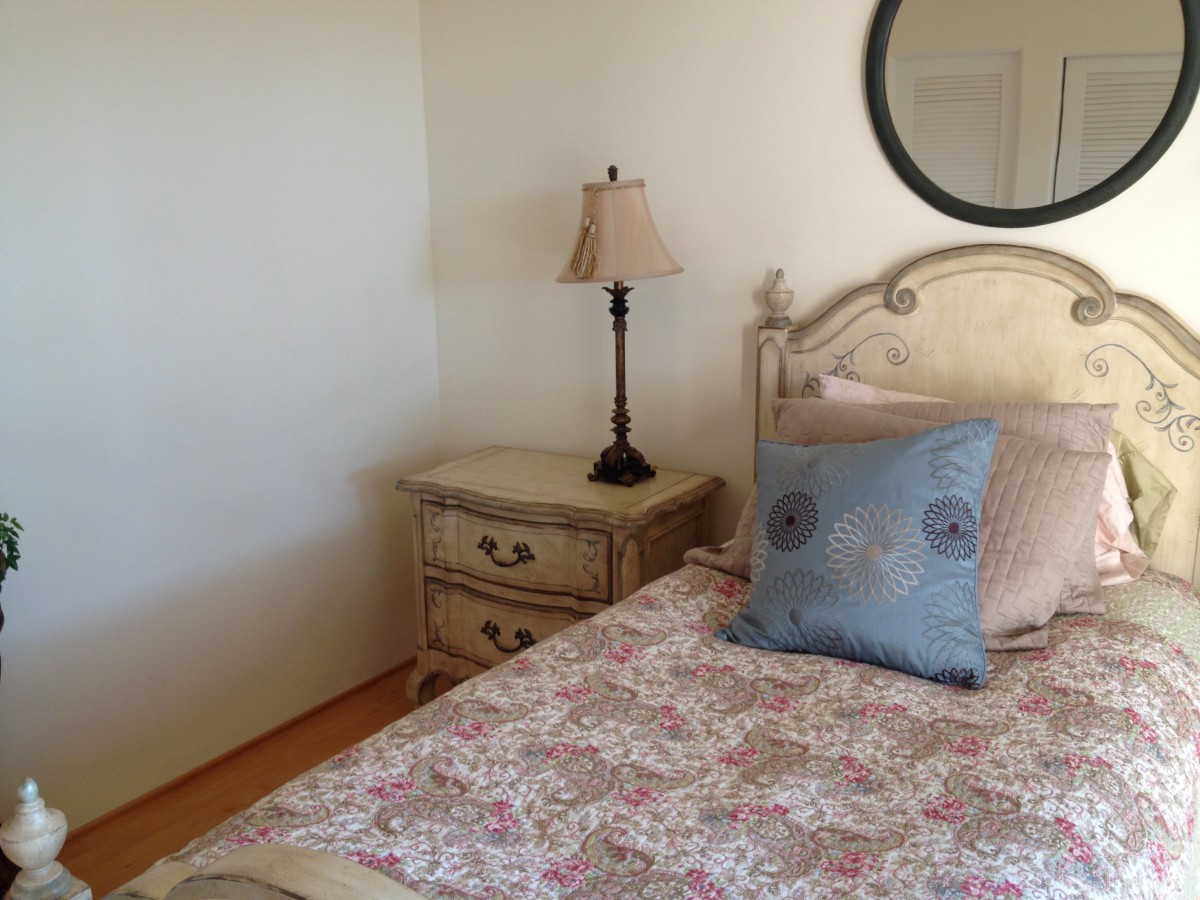 A cozy bedroom with a floral bedspread, decorative pillow, and a nightstand featuring a lamp and mirror.