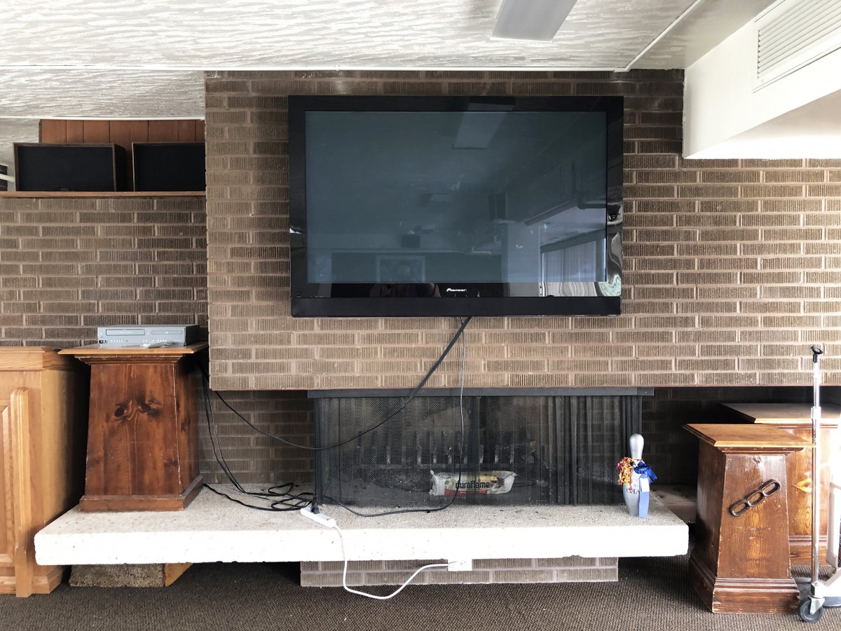 A living room with a television mounted above a brick fireplace, surrounded by various electronics and decor.