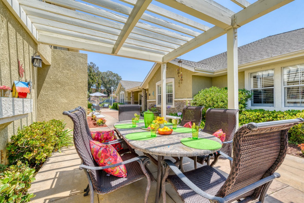 A bright outdoor patio dining area under a pergola, featuring a table set with colorful place settings and surrounded by comfortable chairs.
