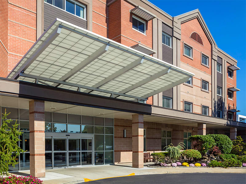 Entrance of a modern hotel building with a large glass awning and landscaped surroundings.