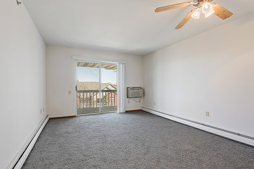 A spacious, empty living room featuring carpet flooring and a ceiling fan, with natural light streaming in from the balcony doors.