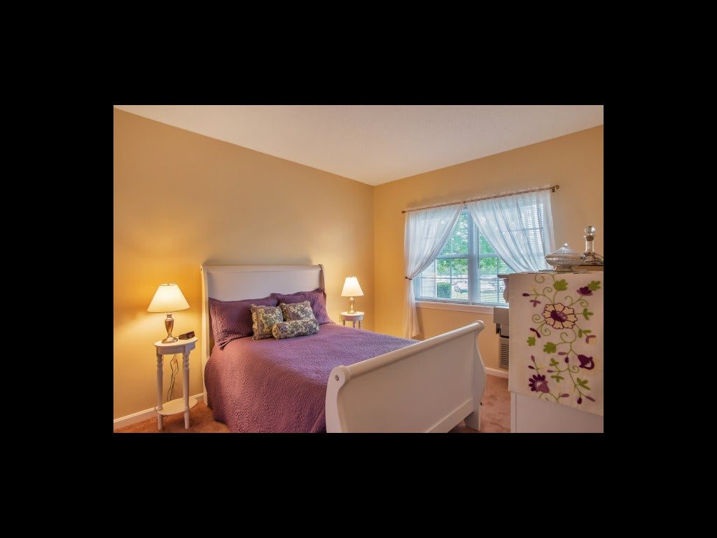 A cozy bedroom with a purple bedspread, side tables with lamps, and a dresser near a window.