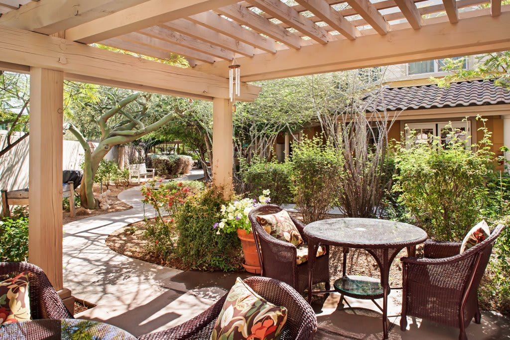 A cozy outdoor patio with comfortable seating surrounded by lush greenery and a wooden pergola overhead.