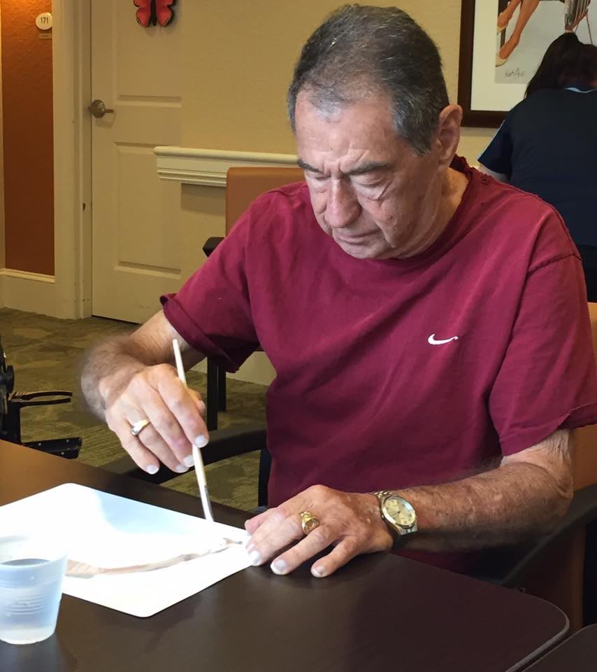 An elderly man in a red shirt is writing on paper at a table indoors.
