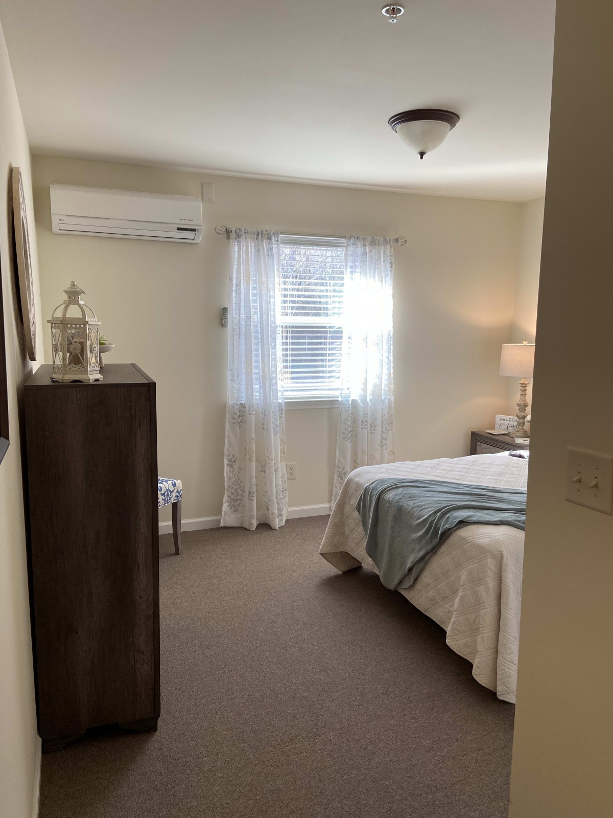 A cozy bedroom with a neatly made bed, dresser, and window with sheer curtains.