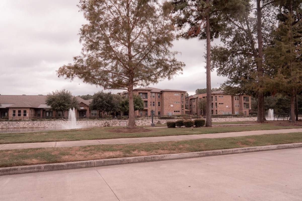 A scenic residential area with apartments by a lake, featuring a fountain and trees under an overcast sky.