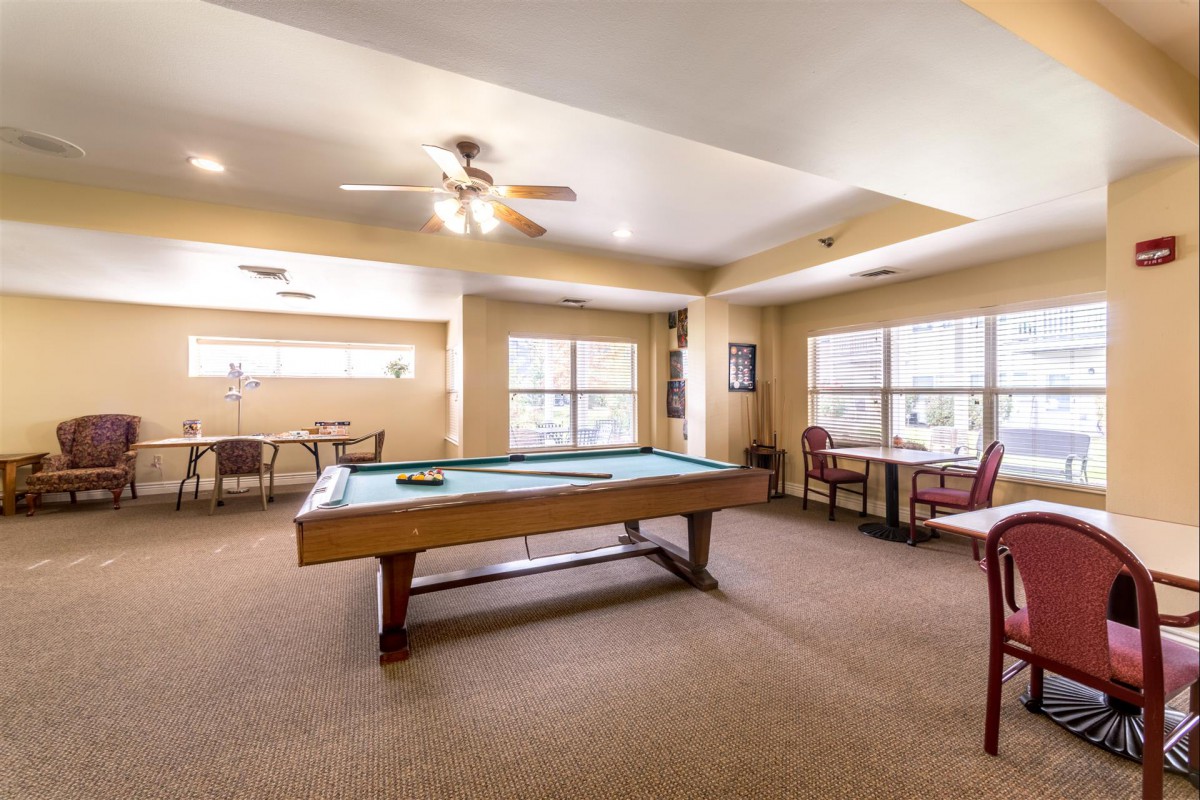 A spacious game room featuring a pool table surrounded by chairs and tables, lit by natural light through large windows.