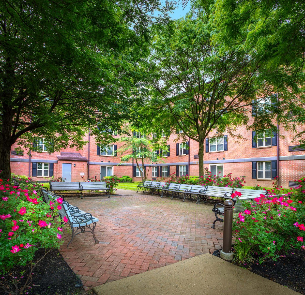 A peaceful courtyard with benches surrounded by lush greenery and a brick building.