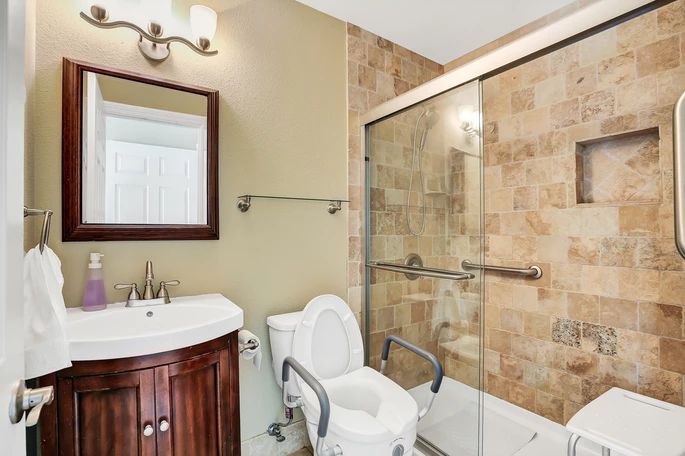 A modern bathroom with a toilet equipped with support rails, a sink with a mirror above, and a shower enclosed by glass doors.