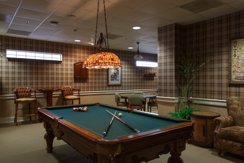 A cozy billiard room featuring a pool table under a decorative hanging lamp, surrounded by warm wood paneling and comfortable seating.