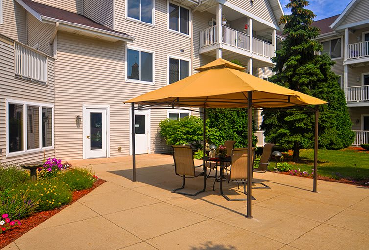 A sunny patio area with outdoor furniture and a large umbrella, adjacent to a building and surrounded by garden landscaping.
