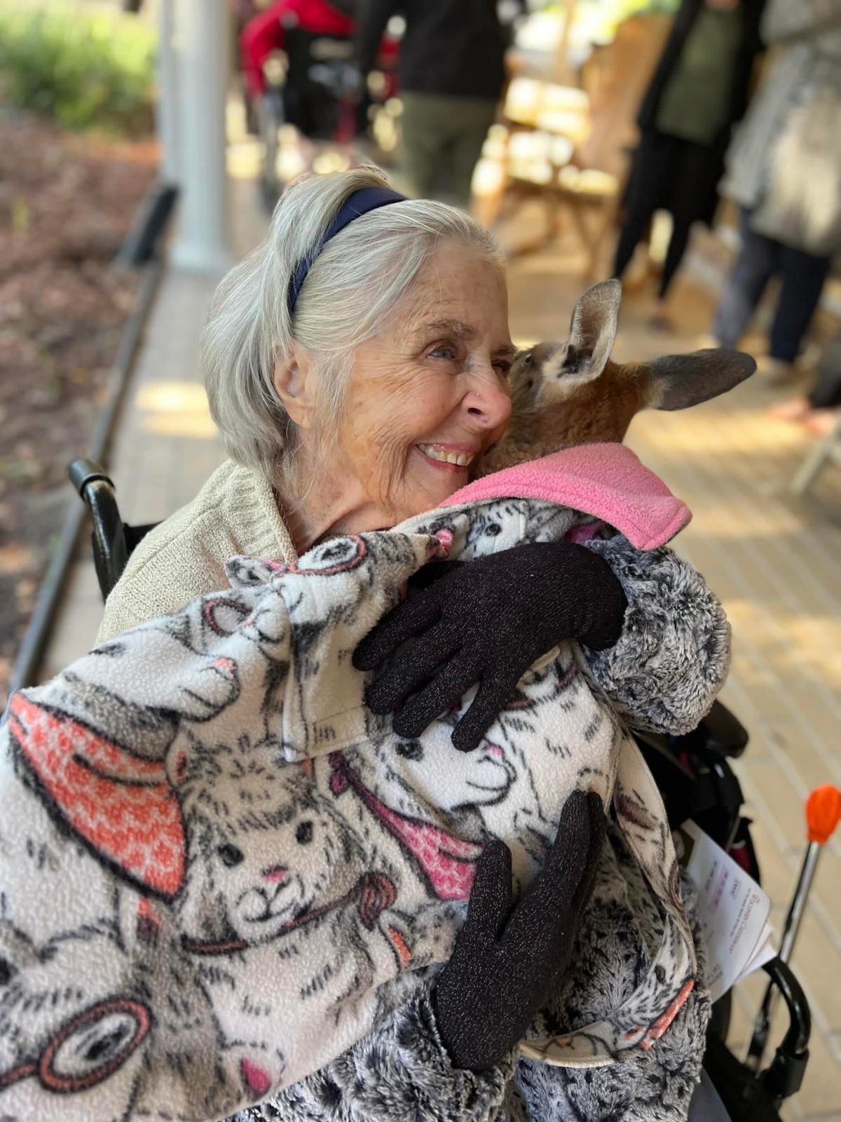 An elderly woman sitting in a wheelchair is joyfully hugging a small goat wrapped in a blanket outdoors.