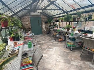 A cozy greenhouse interior filled with plants, tables, and chairs under natural light.