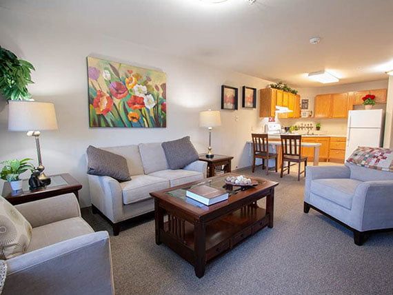 A cozy living room with a sofa set and coffee table, featuring colorful artwork on the wall and an open view of the kitchen area.