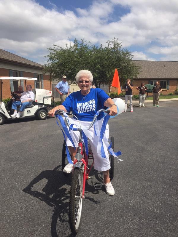 An elderly woman wearing a blue t-shirt rides a bicycle adorned with ribbons during an outdoor celebration.
