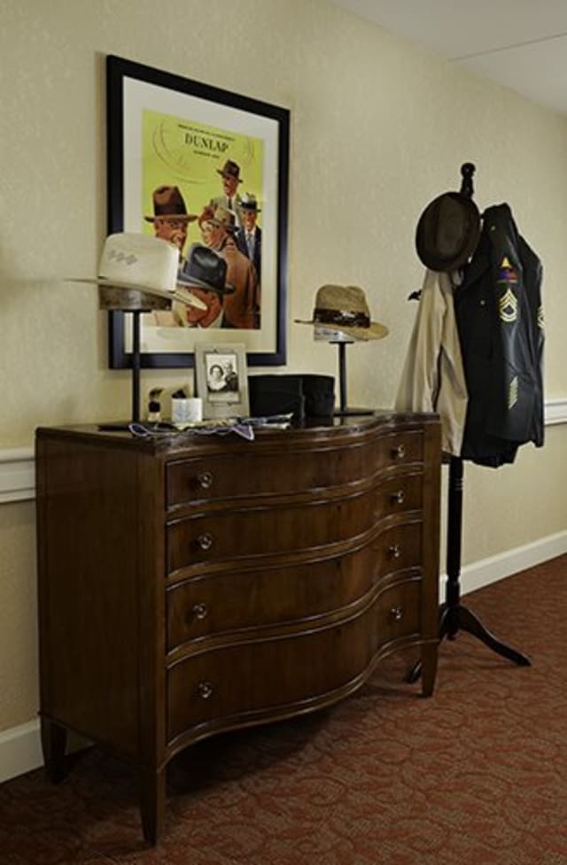 A cozy corner featuring a wooden dresser topped with hats and photographs, next to a coat rack holding jackets and a hat.