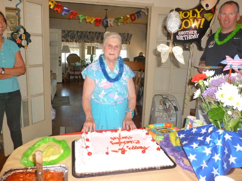 An elderly woman stands in front of a birthday cake with party decorations around her.