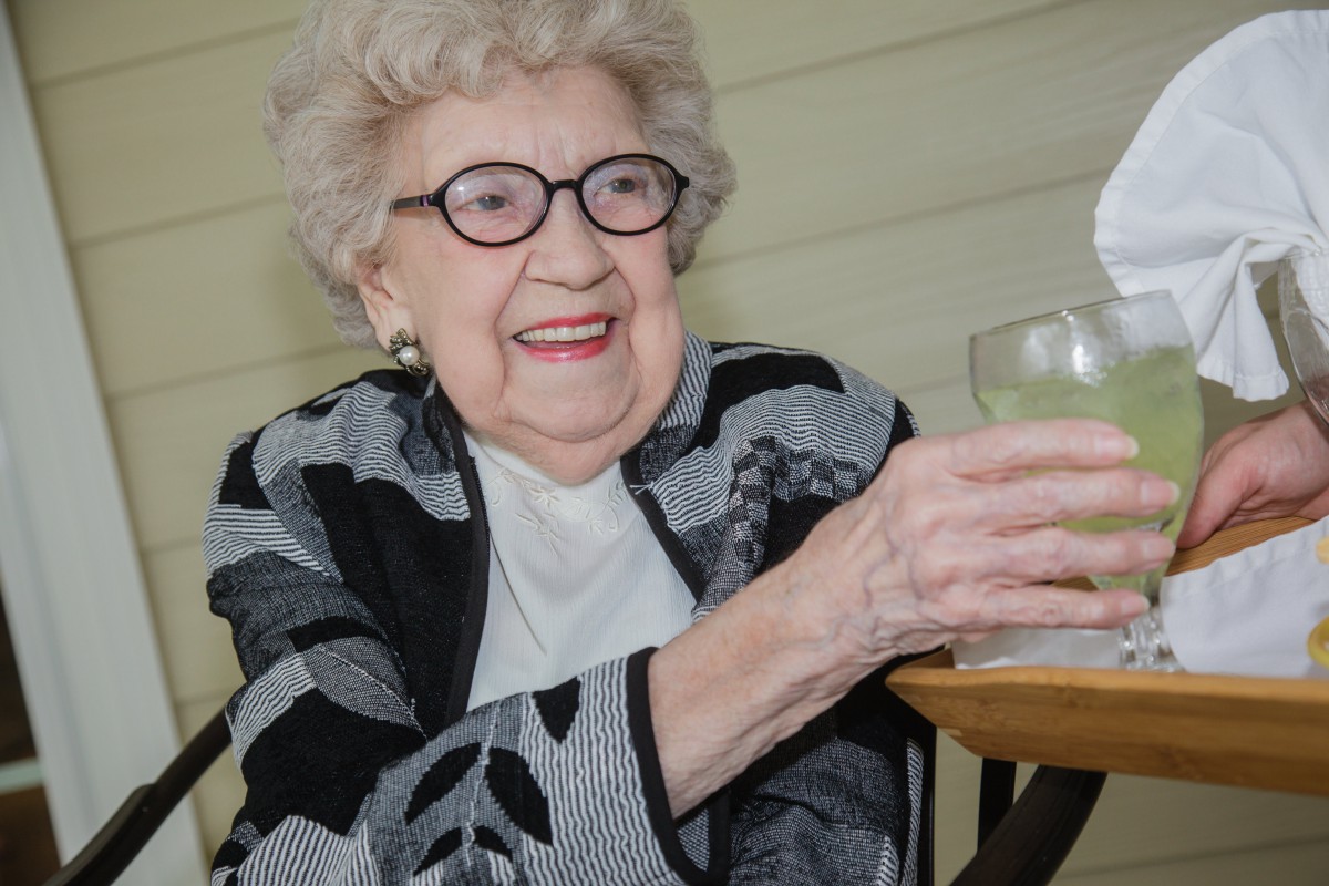 An elderly woman smiling and holding a drink indoors.