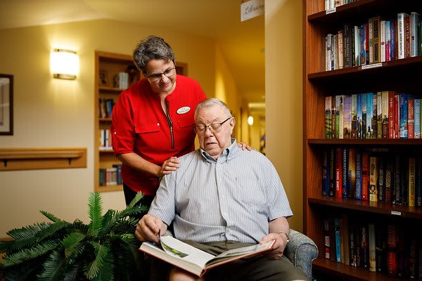 An elderly man reading a book with the assistance of a caregiver in a library setting.