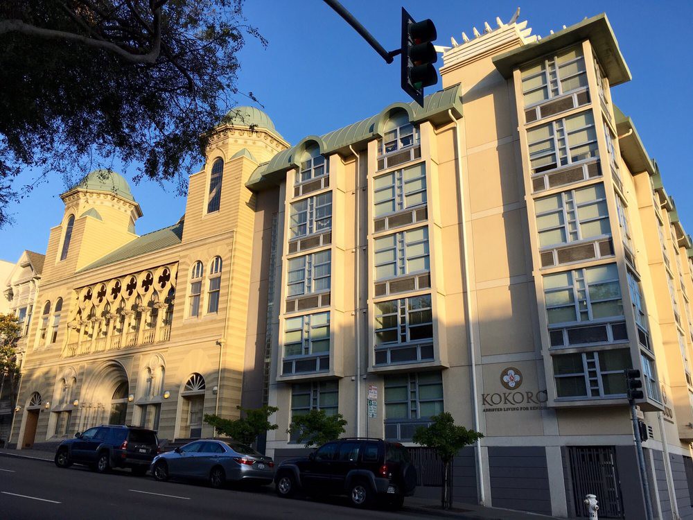 A street view of a multi-story building with distinctive architecture, featuring large windows and cars parked along the roadside.