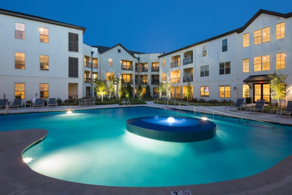 A well-lit apartment complex with a central swimming pool during the evening.