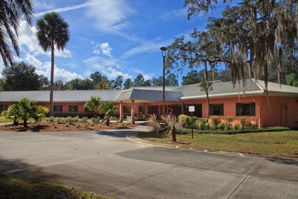 A single-story brick building surrounded by landscaped greenery and trees, with a parking area in front under a blue sky with scattered clouds.