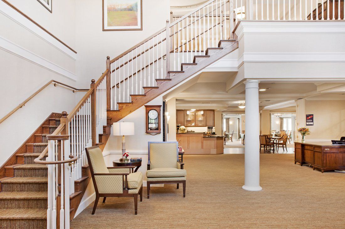 A spacious and well-lit lobby featuring a wooden staircase, comfortable seating area with two chairs and a table, and a view into an adjoining room.