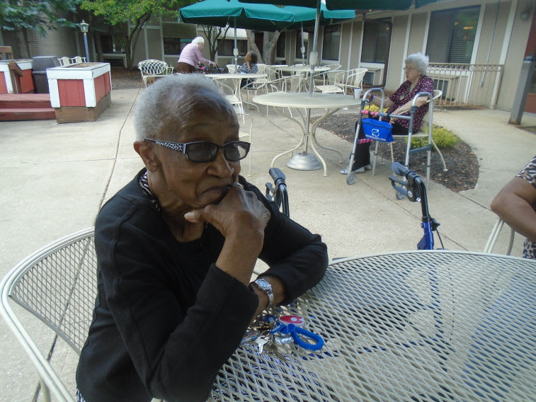 An elderly woman sitting at an outdoor table, looking thoughtful on a sunny day.