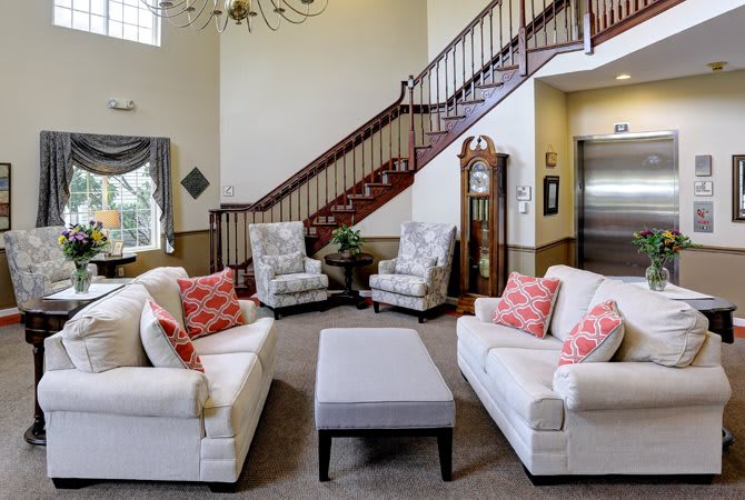 A cozy living room with white sofas and armchairs, decorated with pink cushions, centered around a coffee table, and featuring a grand staircase in the background.