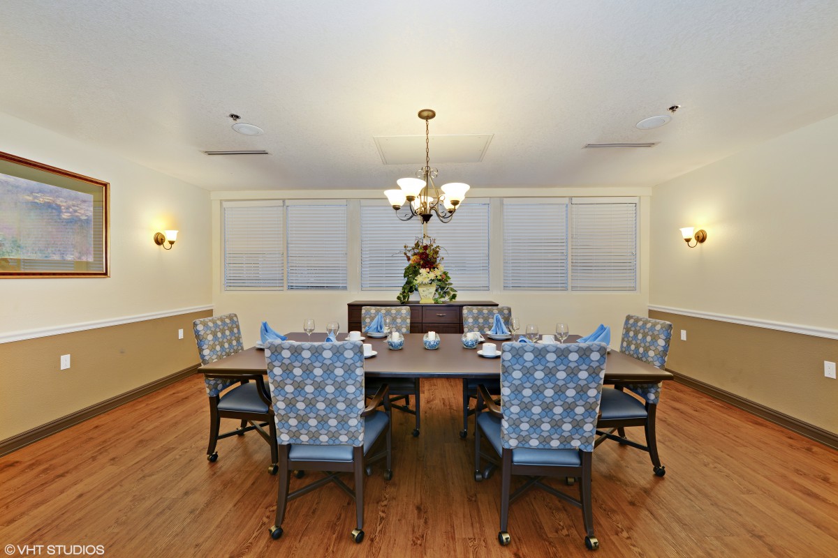 A well-lit dining room with a large table, surrounded by patterned chairs, featuring a central chandelier and wooden flooring.