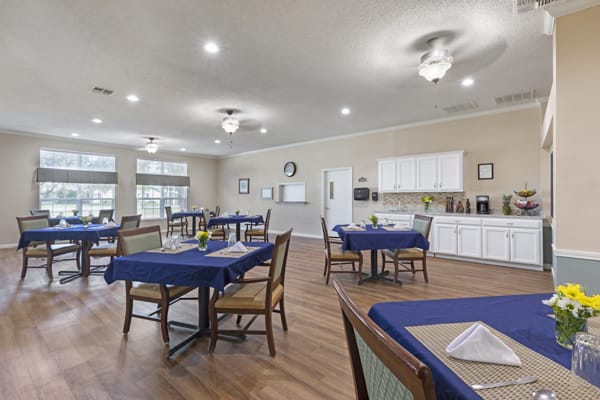 A spacious dining area with wooden floors and tables covered in blue tablecloths, adjacent to a kitchen.