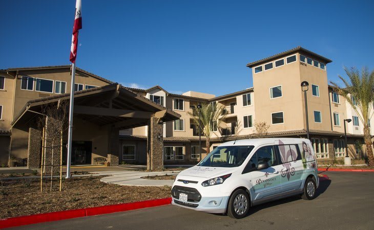 A white van parked in front of a multi-story building with an American flag nearby.