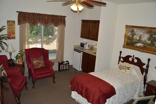 A cozy bedroom featuring a bed, a comfortable chair, and a kitchenette area, illuminated by natural light from the window.