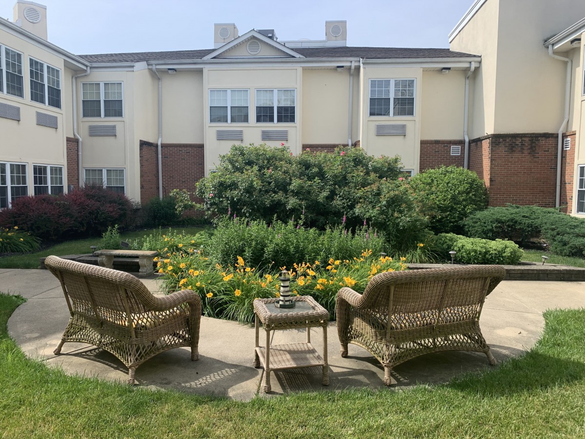 A serene garden seating area with wicker chairs and lush greenery in front of a residential building.