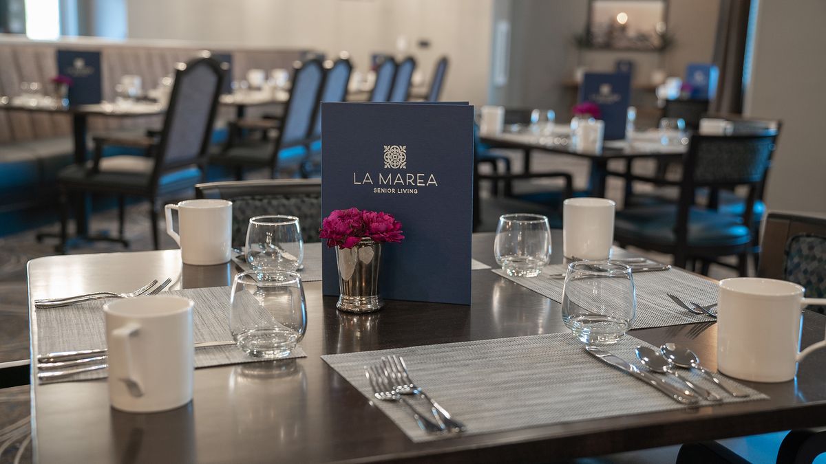 A neatly set dining table at a restaurant with a menu, glasses, cutlery, and a small vase of flowers.