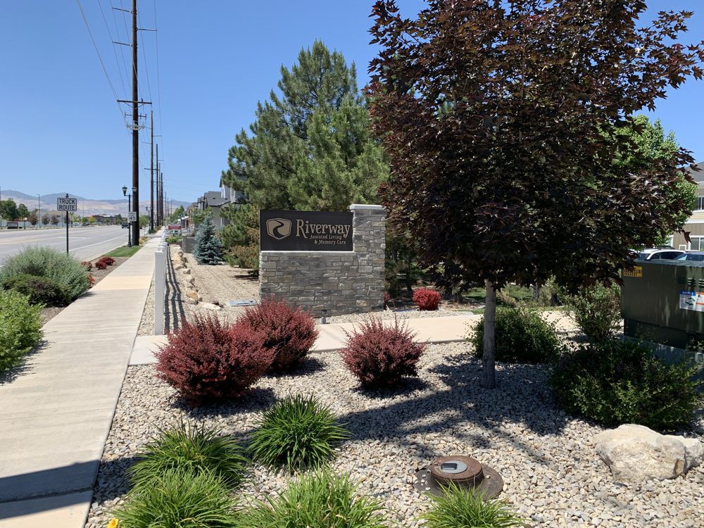 An entrance sign with surrounding landscaping including bushes and trees along a sidewalk.