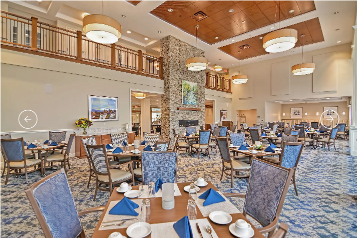 A spacious and elegantly furnished dining room with neatly arranged tables and chairs, featuring a central stone fireplace and stylish chandeliers.
