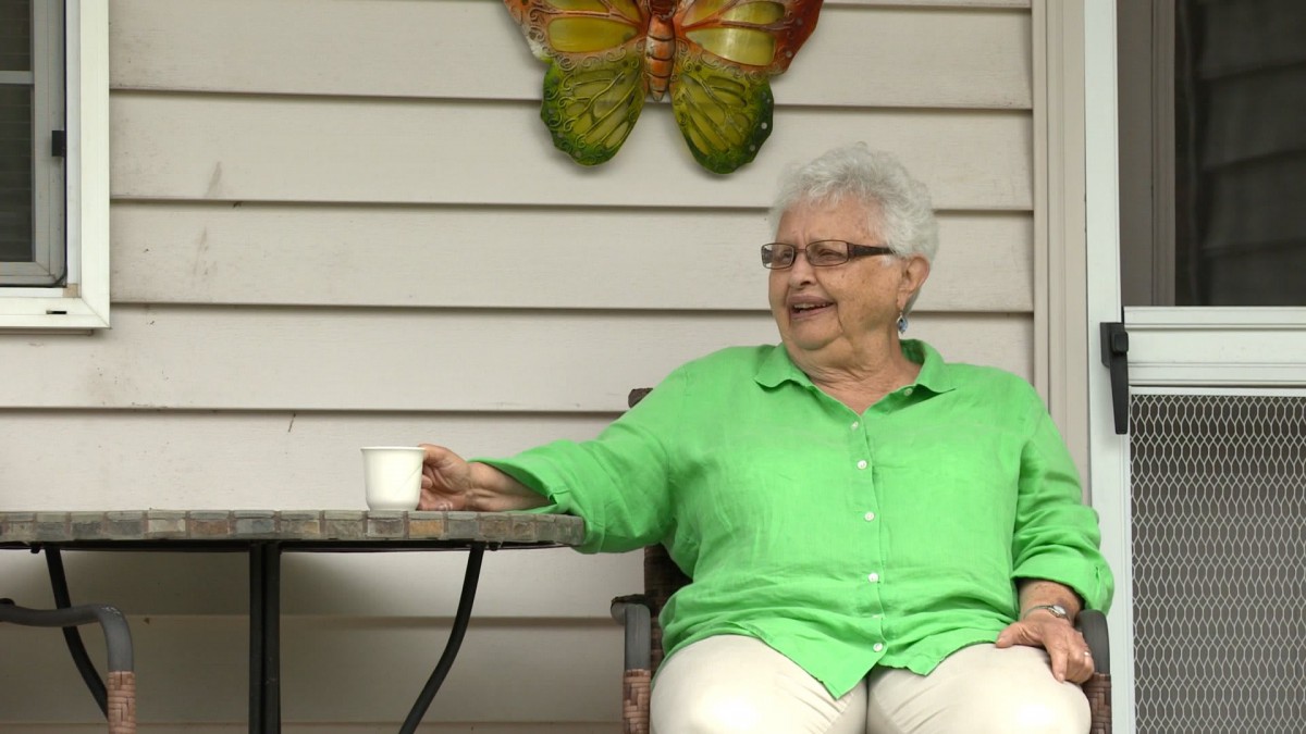 An elderly woman in a green shirt sits outside with a coffee cup, smiling next to a butterfly decoration.