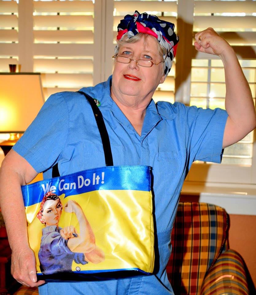 An elderly woman poses indoors with a Rosie the Riveter-themed bag, flexing her arm in a strong pose.