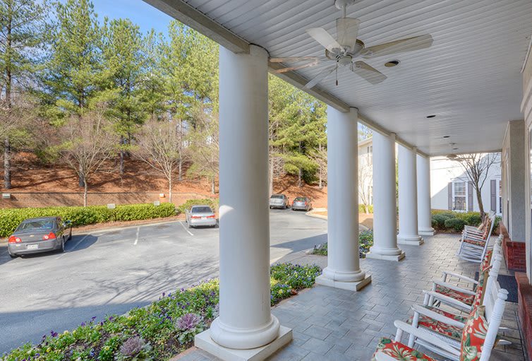 A porch with large columns overlooking a driveway and parked cars, surrounded by trees.