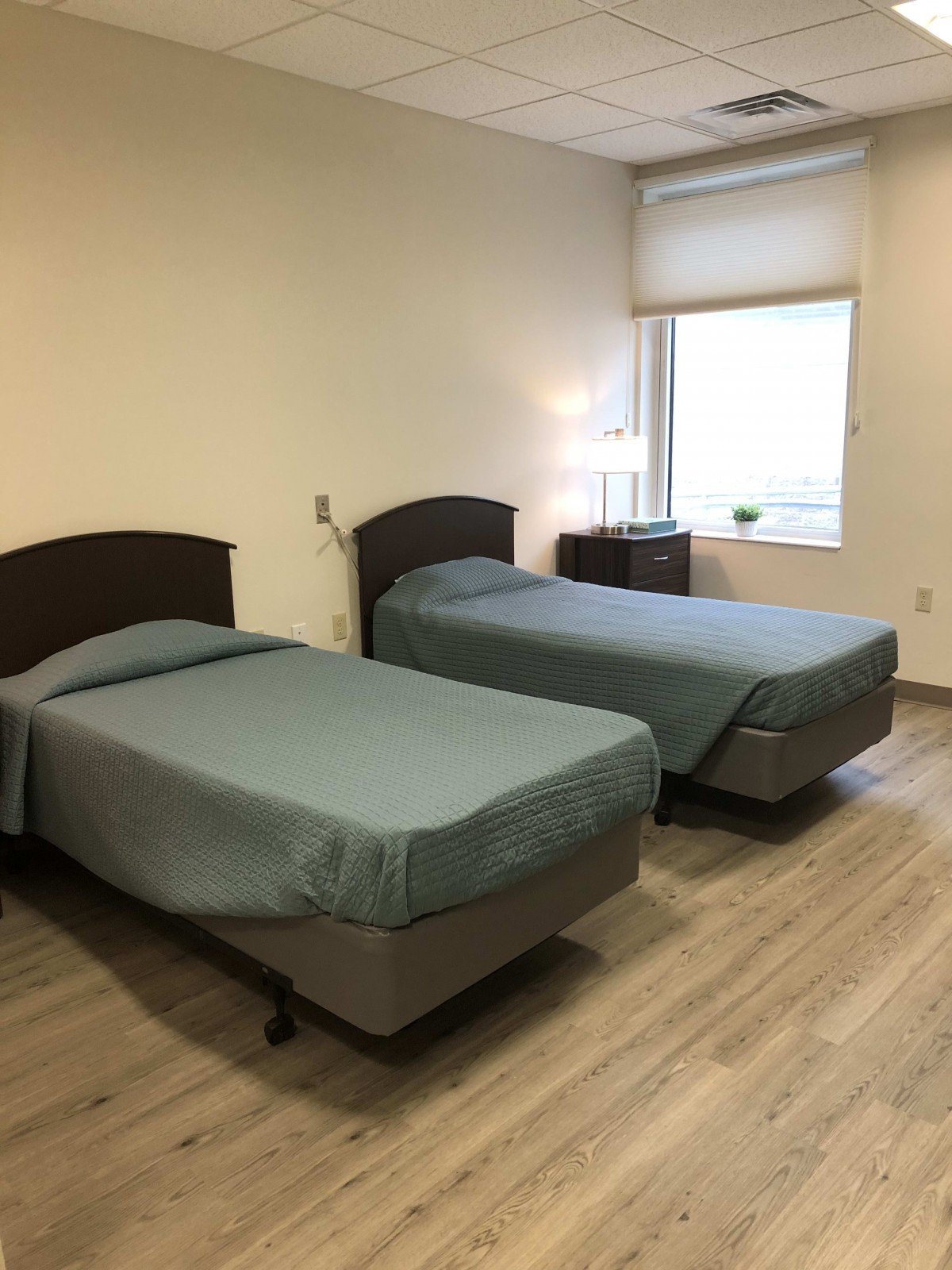 A minimalist hospital room with two neatly made twin beds and light wood flooring.