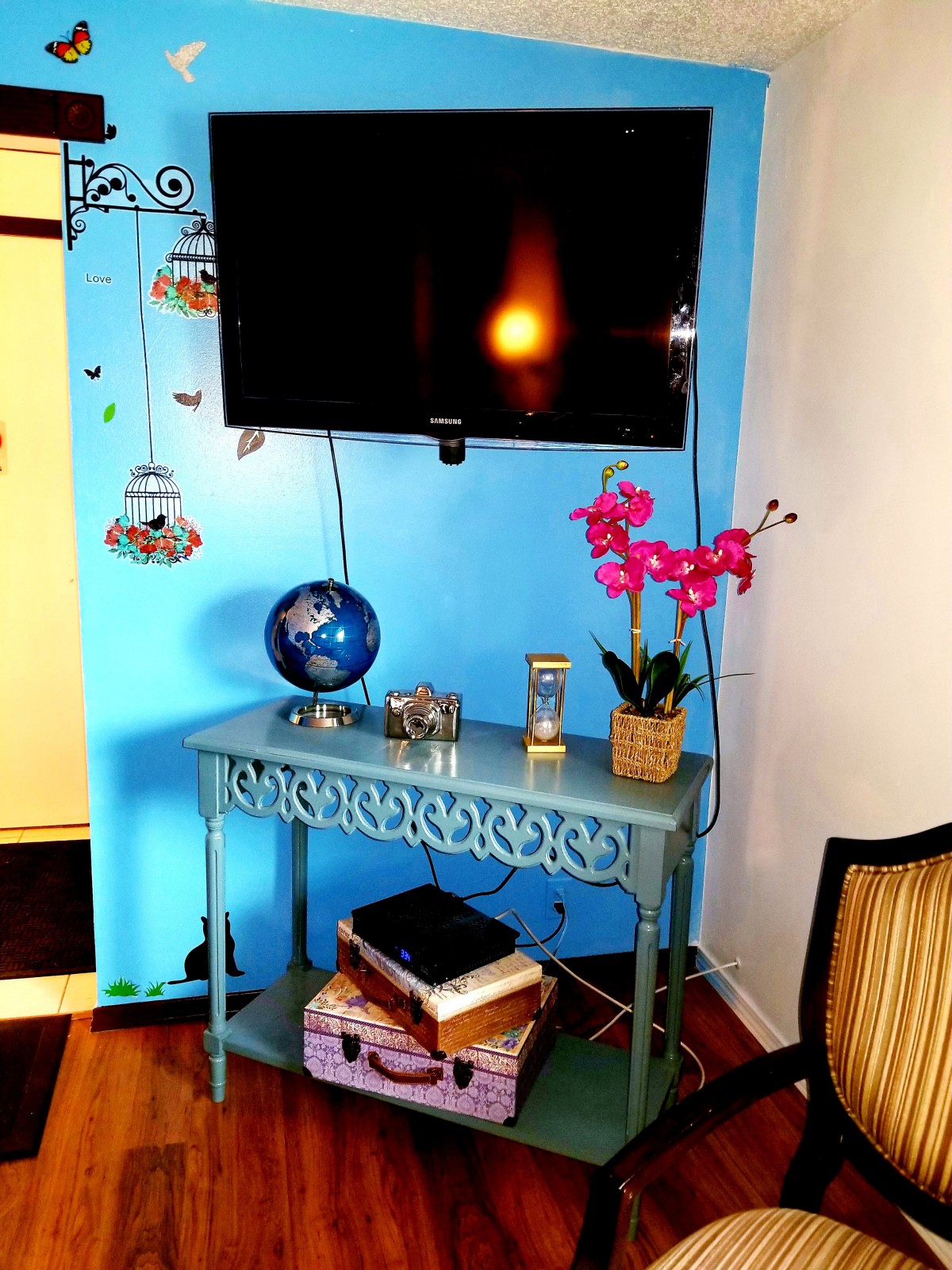 A cozy living room corner featuring a wall-mounted TV, decorative table with a globe, books, and a vibrant orchid.