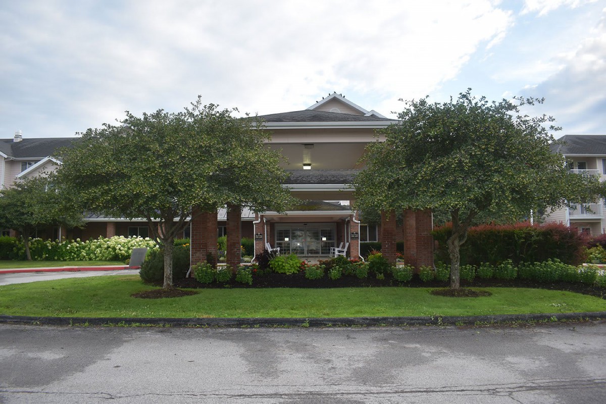 A brick building with a covered entrance, surrounded by landscaped greenery and trees under a partly cloudy sky.
